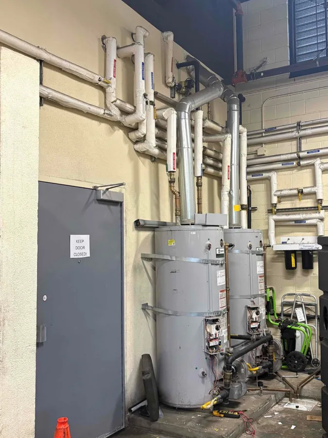 Two commercial tank water heaters side by side with vent pipes and insulated piping running up the wall of a mechanical room