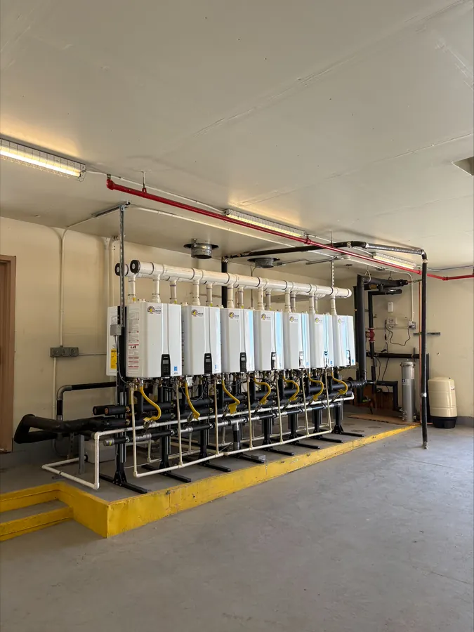 Seven tankless water heaters mounted side by side on a steel rack above a yellow-painted platform, with PVC venting overhead