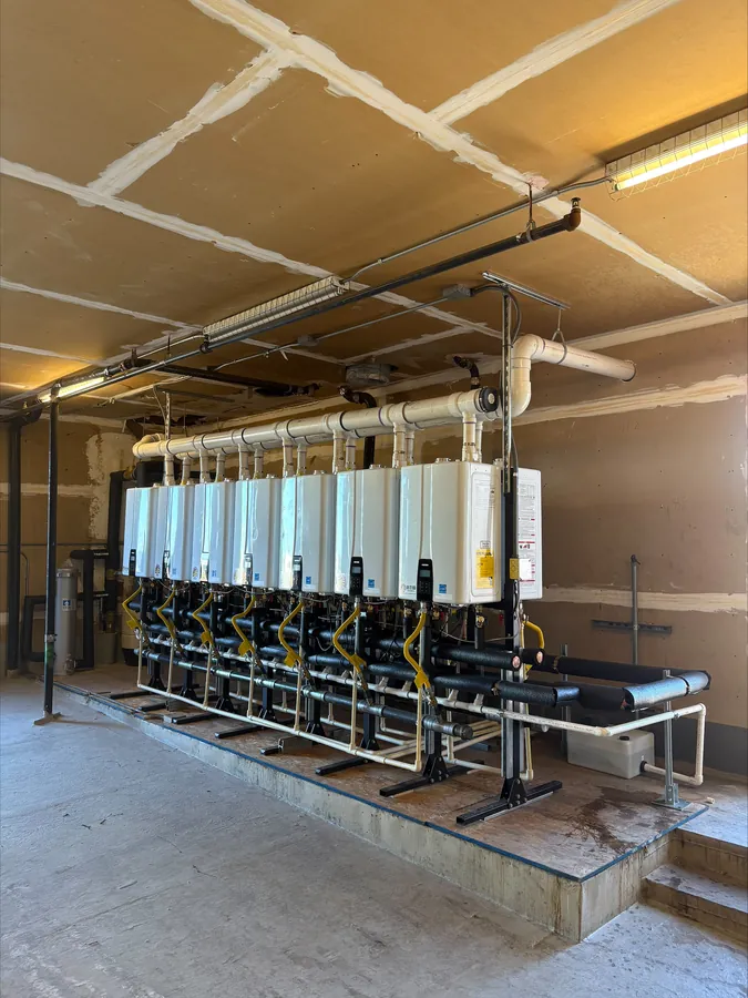 A bank of nine tankless water heaters mounted on a steel frame over a raised concrete platform, with venting and gas lines overhead