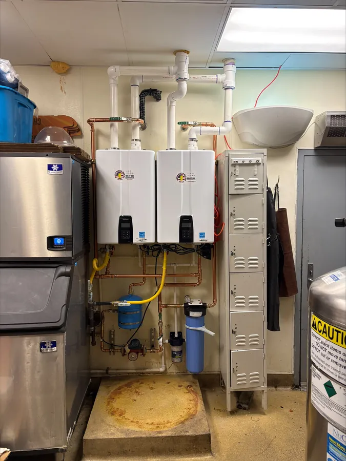 Two wall-mounted tankless water heaters with copper and yellow gas piping, next to an ice machine and storage lockers in a commercial back room