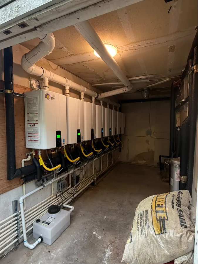 A long row of wall-mounted tankless water heaters with gas and water piping along a brick wall in a commercial utility space