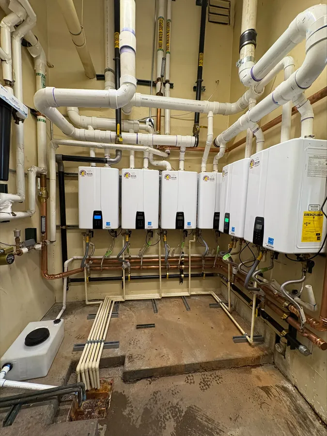 Six tankless water heaters with white PVC vent pipes above and copper water piping below, one unit showing an EnergyGuide label