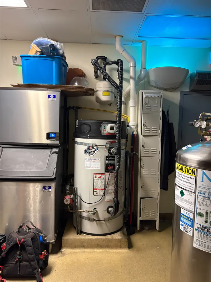 A gas tank water heater with its vent and gas connection in a commercial back room, between an ice machine and storage lockers